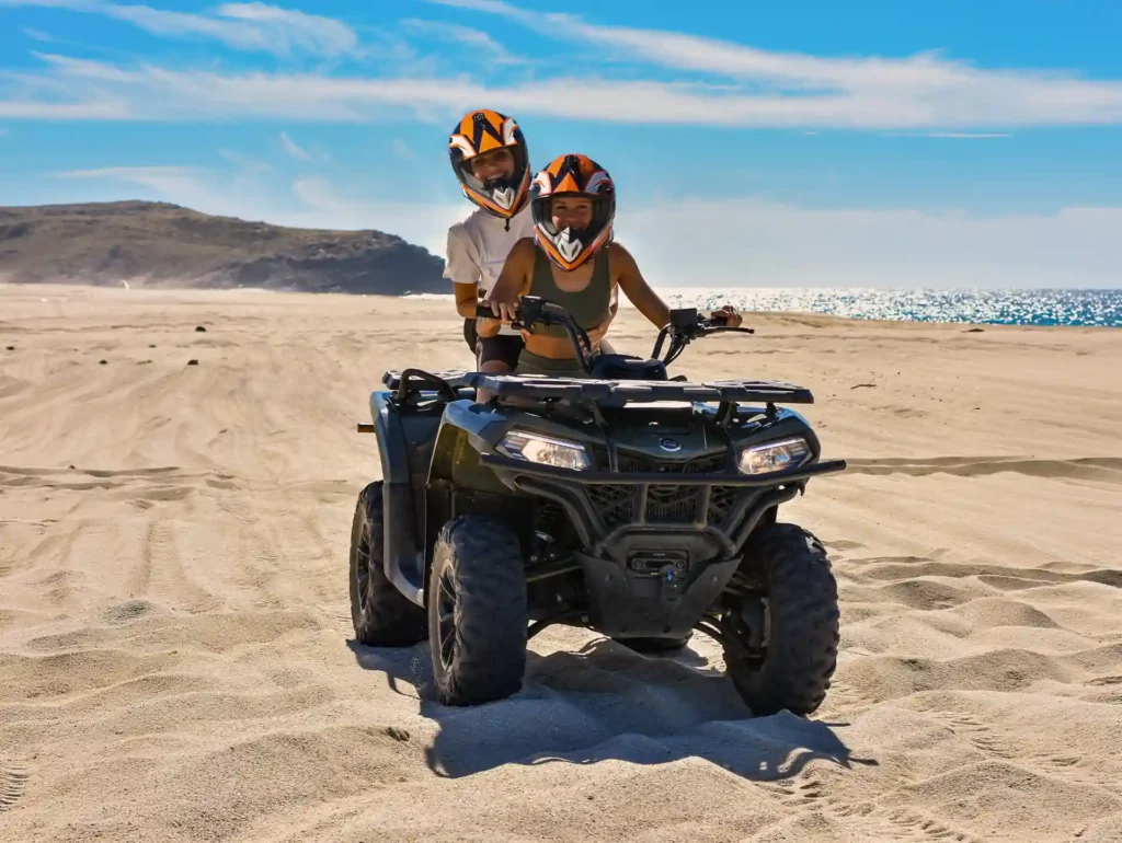 Two people wearing helmets riding an automatic ATV on the sand along a beach with an ocean view.