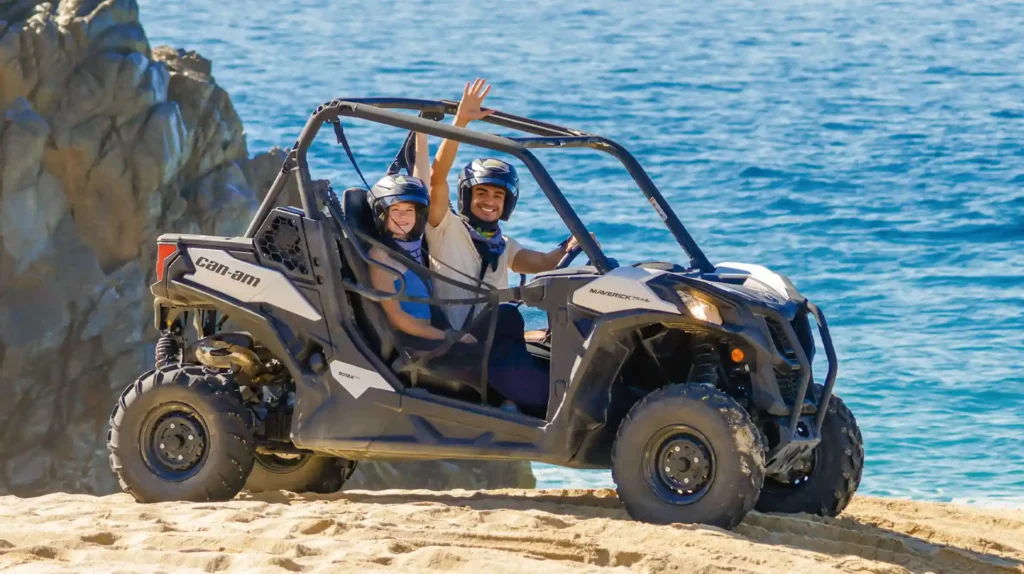 Couple waving while driving a Side by Side UTV next to the ocean in Los Cabos