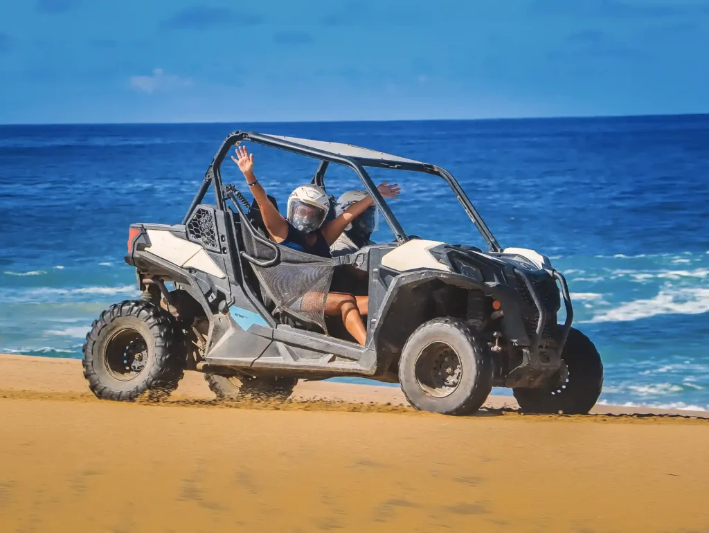 People driving a Side by Side vehicle on the sand by the ocean, with waves in the background.