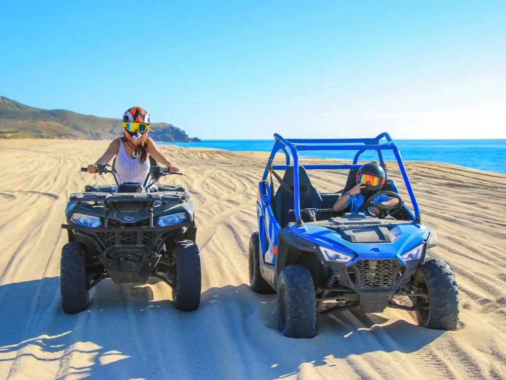 ATV y mini RZR conduciendo sobre la arena en la playa, con vista al mar y dunas al fondo.