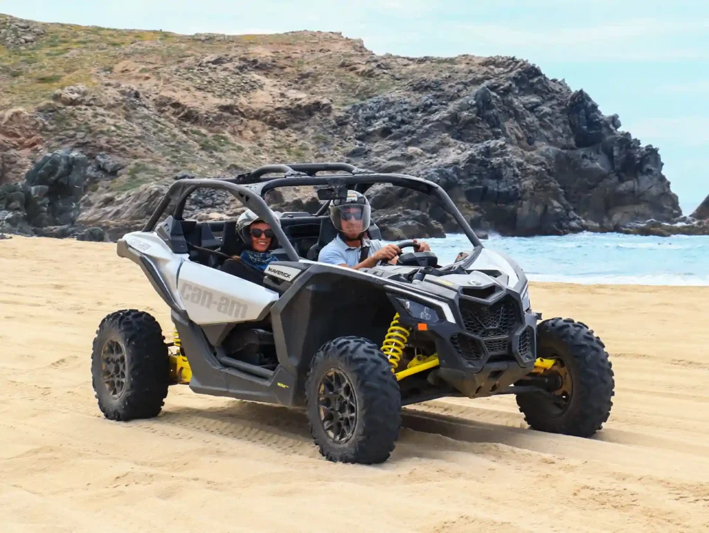 Couple driving a Maverick X3 UTV on the beach sand with rocky formations in the background.