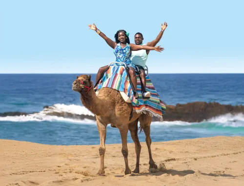 Couple riding a camel on the sand by the ocean during a beach camel tour.